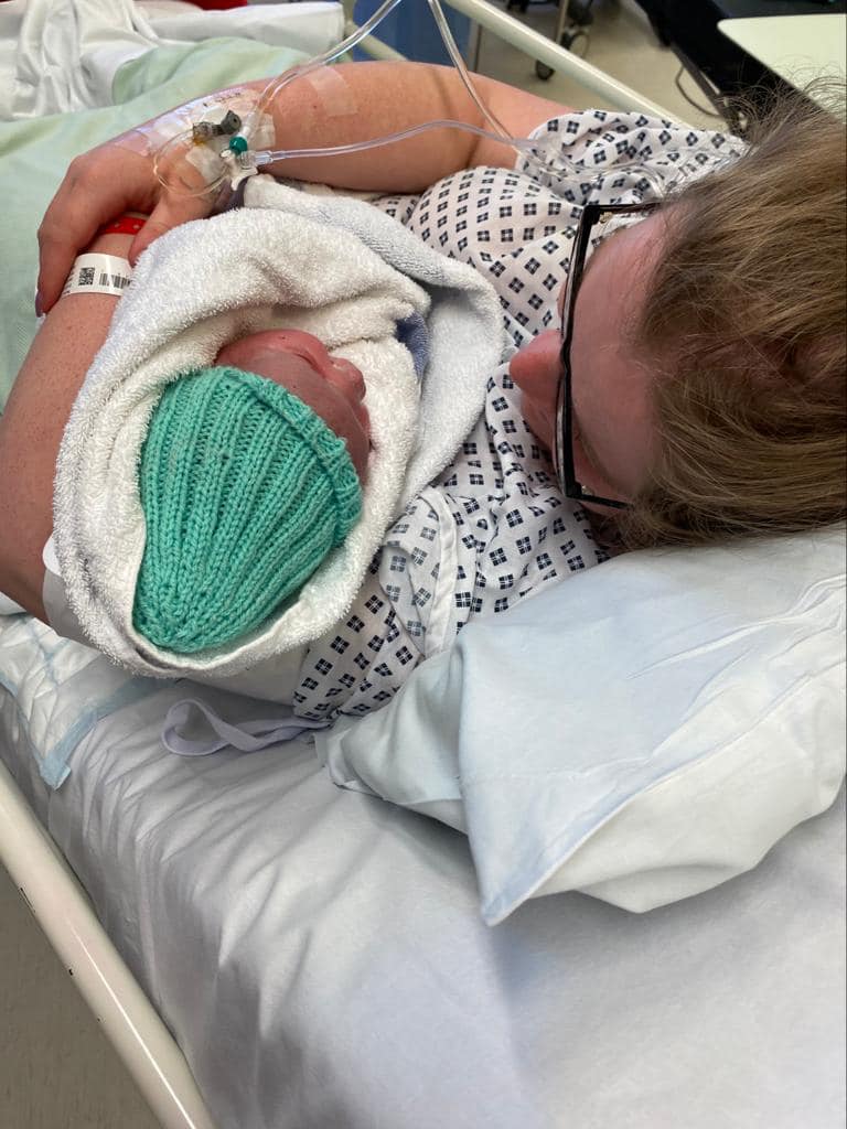 Photo taken from behind and above, Flo is lying on a hospital bed cradling her newborn after a C-Section birth. Baby is wrapped in a towel wearing a green hat. 
