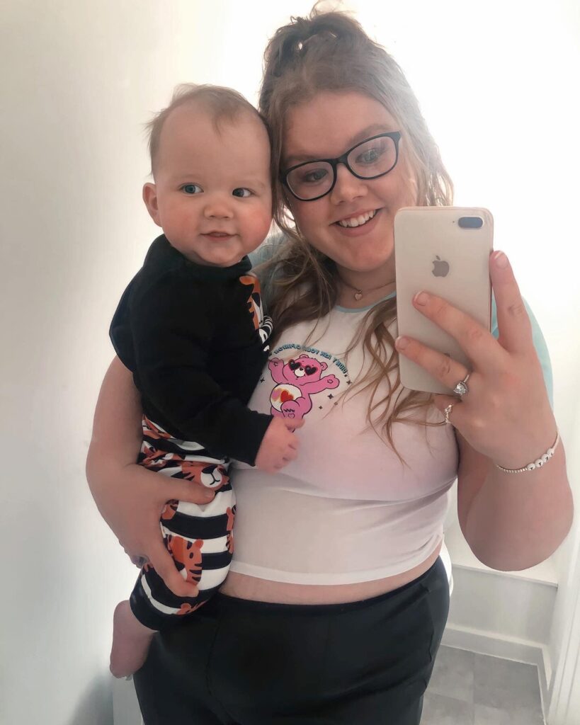 motherhood, Flo holds her son Jett. She is taking a selfie in the mirror with her phone. Her and Jett arr smiling into the mirror.